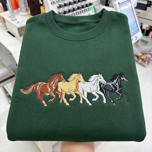 May include: A forest green crewneck sweatshirt featuring an embroidered design of four horses running in a line. The horses are depicted in brown, tan, white, and black thread, showcasing detailed manes, tails, and muscular forms on the fabric.