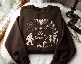 May include: A dark brown crewneck sweatshirt featuring a cream-colored graphic print with the text "respect the locals" surrounded by illustrations of a moth, a UFO, a ghost, a Bigfoot, a sea monster, an alien, and a skeletal deer creature.