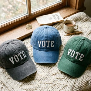May include: Three baseball caps in charcoal gray, denim blue, and forest green, each featuring the word VOTE embroidered in white capital letters on the front panel. The caps have a structured crown, a curved brim, and a distressed fabric finish.