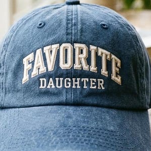 May include: A blue baseball cap with a distressed, washed-out finish. The front features embroidered text in a cream-colored, block-style font that reads FAVORITE in an arched design above the word DAUGHTER in a straight, horizontal line.