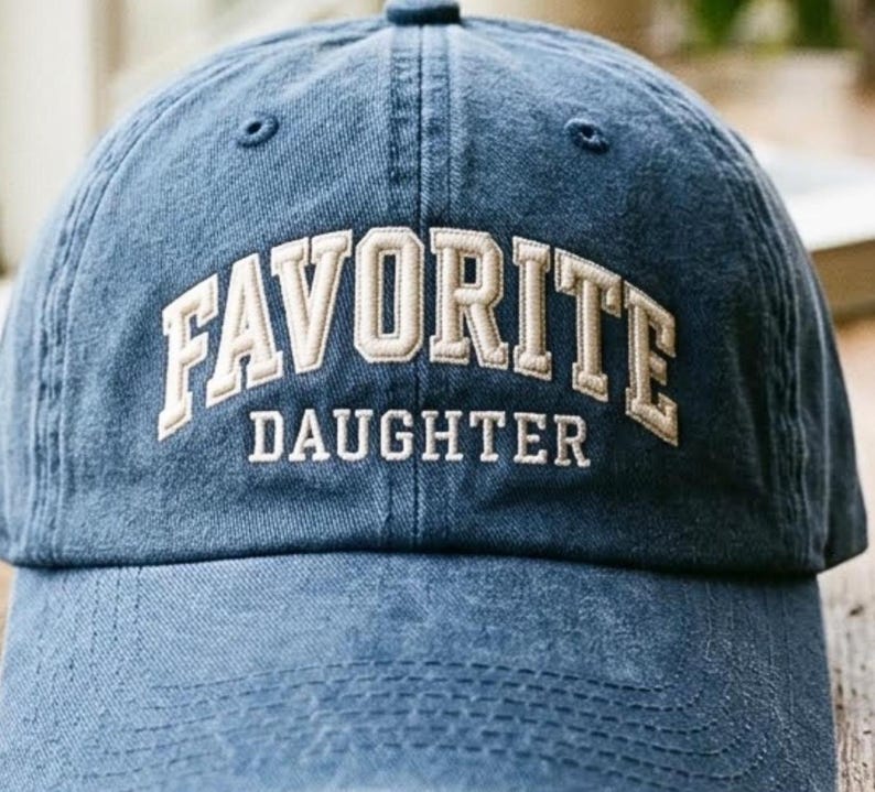May include: A blue baseball cap with a distressed, washed-out finish. The front features embroidered text in a cream-colored, block-style font that reads FAVORITE in an arched design above the word DAUGHTER in a straight, horizontal line.