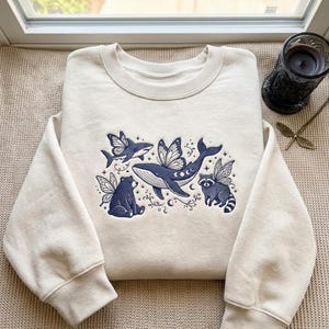 May include: A cream-colored crewneck sweatshirt featuring dark blue embroidery of a whale, a shark, a bear, and a raccoon, all depicted with butterfly wings. The design includes small stars, crescent moons, and swirling decorative line elements.