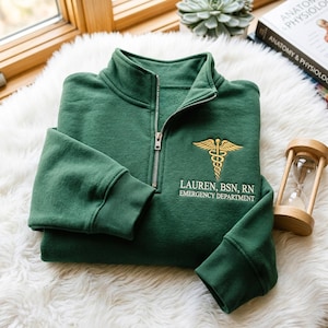 Pode incluir: Uma camisola verde-escura com fecho de correr curto e um símbolo de caduceu dourado bordado no lado esquerdo do peito. Abaixo do símbolo, o texto LAUREN, BSN, RN e EMERGENCY DEPARTMENT está escrito em branco. Mangas compridas com punhos canelados.
