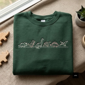 Op de afbeelding: Een donker bosgroen sweatshirt met ronde hals en een rij geborduurde dinosaurussen in lichtroze draad. Het ontwerp bevat een triceratops, een theropode, een langhals-sauropode, een pterosauriër, een stegosaurus en een ander vliegend reptiel.