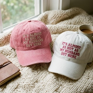 May include: A pink baseball cap with white embroidered text reading IT'S ALMOST AS HOT AS MY HUSBAND OUT HERE and a white baseball cap with pink embroidered text reading IT'S ALMOST AS HOT AS MY HUSBAND OUT HERE, both featuring a curved brim.