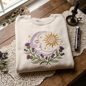 Puede incluir: Una sudadera de cuello redondo color crema con un diseño bordado de una luna creciente lavanda con rostro, un sol dorado y plantas de cardo verdes con flores moradas. El bordado está centrado en la parte frontal de la prenda.