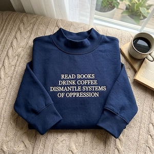 May include: A navy blue long-sleeve sweatshirt with a mock neck collar. The front features cream-colored embroidered text that reads: READ BOOKS DRINK COFFEE DISMANTLE SYSTEMS OF OPPRESSION. The garment has ribbed cuffs and a hem.