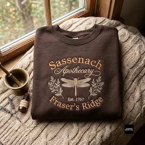 Puede incluir: Una sudadera de cuello redondo color marrón oscuro con texto bordado en dorado que dice Sassenach Apothecary Est. 1767 Fraser's Ridge. El diseño incluye una libélula central flanqueada por dos ramas con hojas, centrada en el pecho.