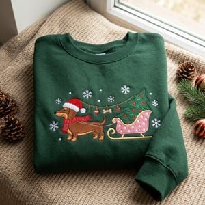 May include: A dark green crewneck sweatshirt featuring an embroidered design of a brown dachshund wearing a red Santa hat and scarf, pulling a pink polka-dot sleigh carrying a green Christmas tree, surrounded by white embroidered snowflakes.