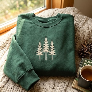 Peut inclure: Un sweat-shirt à col rond vert forêt arborant une broderie centrale de couleur crème représentant trois pins. Le vêtement est doté de manches longues avec des poignets côtelés et d'un col côtelé, offrant une esthétique épurée et sobre.
