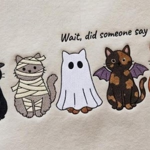 May include: A cream-colored fabric featuring an embroidered graphic of five cats in Halloween costumes: a black cat in a witch hat, a gray mummy cat, a white ghost cat, a tortoiseshell bat cat, and an orange pumpkin cat with the text Wait, did someone say treats?