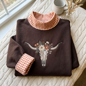 May include: A dark brown long-sleeve sweatshirt featuring a mock neck collar with an orange and white gingham pattern. The cuffs also display the same gingham print. An embroidered steer skull with a floral arrangement is centered.