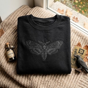 May include: A black crewneck sweatshirt featuring a dark grey embroidered moth design on the chest. The moth has intricate wing patterns and a small skull detail on its thorax. The fabric has a smooth texture and a classic long-sleeve silhouette.