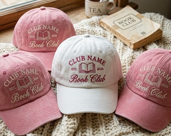May include: Three baseball caps in a group, two in dusty pink and one in white. Each cap features embroidered text reading CLUB NAME, EST. 2026, and Book Club, centered above an open book icon. The embroidery is white on the pink caps and maroon on the white cap.