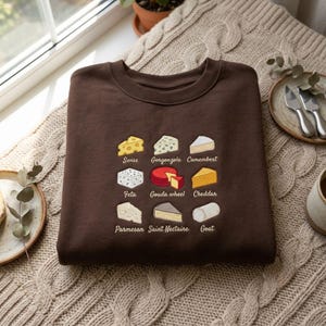 Può includere: Una felpa girocollo marrone scuro con un ricamo raffigurante nove diversi tipi di formaggio disposti in una griglia. Ogni illustrazione di formaggio è etichettata con il suo nome: Swiss, Gorgonzola, Camembert, Feta, Gouda wheel, Cheddar, Parmesan, Saint Nectaire e Goat.