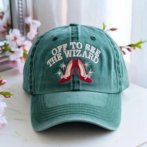 Puede incluir: Una gorra de béisbol verde bosque con texto bordado en blanco que dice OFF TO SEE THE WIZARD. Debajo del texto hay un gráfico bordado de dos zapatos rojos de tacón alto con lazos pequeños, rodeados de detalles de estrellas blancas.