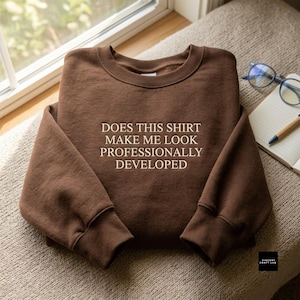 May include: A brown crewneck sweatshirt featuring the embroidered text "DOES THIS SHIRT MAKE ME LOOK PROFESSIONALLY DEVELOPED" in a light beige color. The garment has long sleeves, a ribbed neckline, and ribbed cuffs, presented in a flat lay view.
