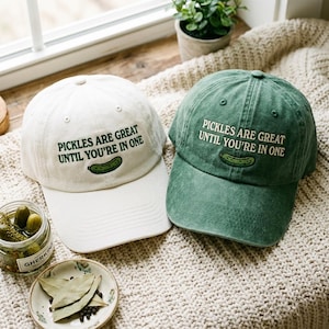 Op de afbeelding: Twee baseballpetten, één gebroken wit en één bosgroen, elk voorzien van donkergroene geborduurde tekst "PICKLES ARE GREAT UNTIL YOU'RE IN ONE" boven een kleine geborduurde groene augurk op het voorpaneel van de pet.