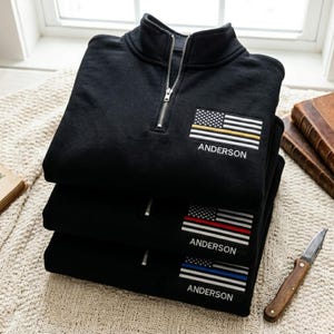 May include: A stack of three black quarter-zip sweatshirts featuring an embroidered American flag patch on the left chest. Each flag includes a single horizontal stripe in gold, red, or blue, with the name ANDERSON embroidered in white below.