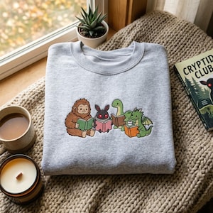 May include: A light gray crewneck sweatshirt featuring an embroidered graphic of a brown Bigfoot, a black moth-like creature with red eyes, a green long-necked dinosaur, and a green dragon, each holding and reading a colorful book.