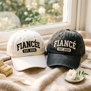 May include: Two baseball caps, one off-white and one charcoal grey, both featuring embroidered text. The off-white cap displays the word FIANCÉE in black, and the charcoal cap displays FIANCÉ in cream, both with the text EST. 2026 below.