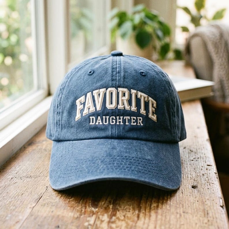 May include: A blue denim baseball cap featuring the embroidered text FAVORITE DAUGHTER in white capital letters across the front panel. The cap has a curved brim, a structured crown with ventilation eyelets, and a classic six-panel construction.