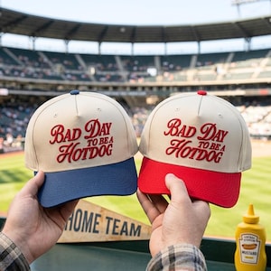 Könnte beinhalten: Zwei Baseballkappen mit beigen Kronen und rotem Sticktext, der Bad Day To Be A Hotdog lautet. Eine Kappe hat einen marineblauen Schirm, die andere einen roten Schirm. Beide Kappen verfügen über ein strukturiertes Design mit fünf Paneelen.