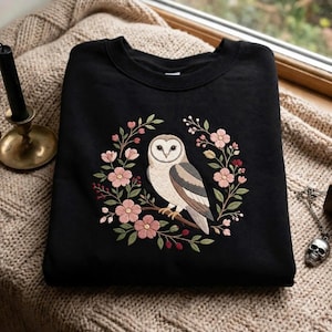 May include: A black crewneck sweatshirt featuring an embroidered design of a barn owl perched on a branch, surrounded by a circular wreath of pink flowers, green leaves, and small red berries. The owl has white, tan, and brown feathers.