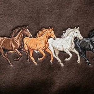 May include: A dark brown sweatshirt featuring an embroidered design of four horses running in a line. The horses are depicted in different colors, including dark brown, light tan, white, and black, with detailed stitching showing their manes.