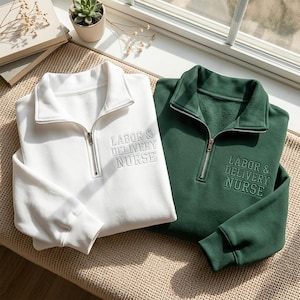 Könnte beinhalten: Ein weißes und ein dunkelgrünes Sweatshirt mit kurzem Reißverschluss, Kragen und langen Ärmeln. Auf der linken Brust beider Kleidungsstücke ist der Text "LABOR & DELIVERY NURSE" in einer zum Stoff passenden Farbe eingestickt.
