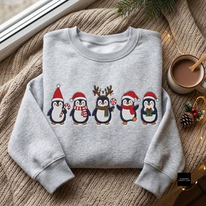 Peut inclure: Un sweat-shirt gris clair &agrave; col rond orn&eacute; d'un motif repr&eacute;sentant cinq pingouins. Les pingouins portent des bonnets de P&egrave;re No&euml;l rouges, des &eacute;charpes et des bois de renne, tout en tenant une sucette, un sucre d'orge et un cadeau vert.