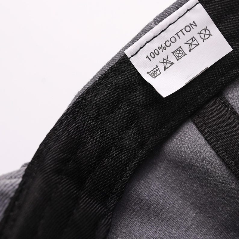 May include: The interior of a grey cap featuring a black fabric sweatband and a white care label. The label is printed with the text 100% COTTON and includes four laundry care symbols indicating a 30 degree wash, no bleaching, no ironing, and no dry cleaning.
