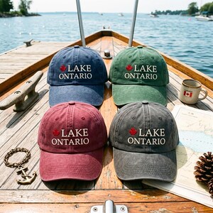 Op de afbeelding: Vier baseballpetten in blauw, groen, bordeauxrood en antracietgrijs, elk voorzien van een geborduurd rood esdoornblad en de witte tekst "LAKE ONTARIO" op de voorzijde. De petten hebben een verwassen afwerking en liggen op een houten bootdek.