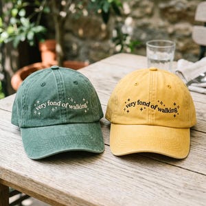 Op de afbeelding: Twee baseballpetten, één in bosgroen en één in mosterdgeel, beide voorzien van de geborduurde tekst "very fond of walking" in een gebogen lettertype omringd door kleine sterpictogrammen op de voorpanelen boven de gebogen kleppen.