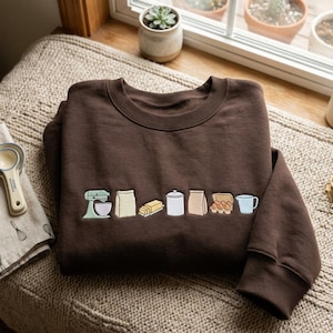 May include: A dark brown crewneck sweatshirt featuring a horizontal row of embroidered baking icons including a light green stand mixer, a bag of flour, a stick of butter, a white canister, a paper bag, a carton of eggs, and a blue pitcher.