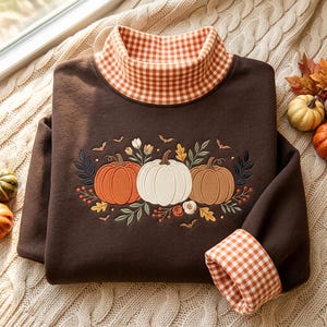May include: A dark brown long-sleeve sweatshirt featuring a high collar and sleeve cuffs with an orange and white gingham pattern. The center of the chest displays an embroidered design of three pumpkins in orange, white, and tan, surrounded by leaves, berries, and small bats.