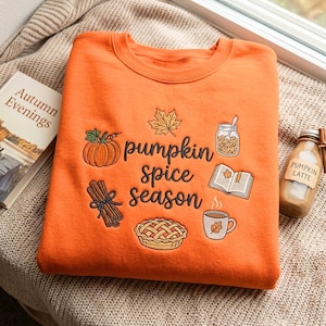 Może przedstawiać: Pomarańczowa bluza z okrągłym dekoltem i ciemnobrązowym haftem z napisem pumpkin spice season. Wzór zawiera ikony dyni, liścia klonu, słoika, otwartej książki, wiązki lasek cynamonu, ciasta oraz kubka z liściem.