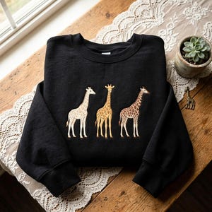 May include: A black long-sleeve crewneck sweatshirt featuring an embroidered design of three giraffes standing side by side. The giraffes are depicted in varying shades of cream, tan, and brown, showcasing their distinct spotted patterns.