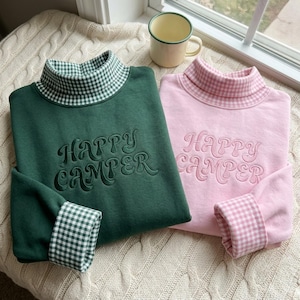 Könnte beinhalten: Zwei langärmelige Sweatshirts, eines in Dunkelgrün und eines in Hellrosa, jeweils mit Stehkragen und Ärmelbündchen in passendem Vichy-Karo-Muster. Der Schriftzug "HAPPY CAMPER" ist in einer geschwungenen Schrift auf der Vorderseite eingestickt.