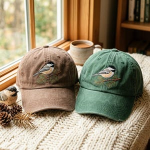 Puede incluir: Dos gorras de béisbol, una marrón y otra verde, cada una con un bordado de un carbonero cabecinegro posado en una rama de pino. Las gorras tienen una corona estructurada, una visera curva y ojales cosidos en los paneles superiores.