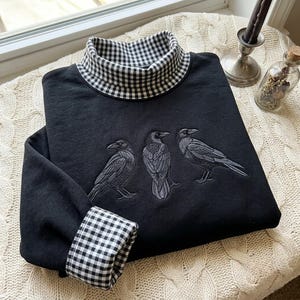 Könnte beinhalten: Ein schwarzes Langarm-Sweatshirt mit Stehkragen und Ärmelbündchen in schwarz-weißem Vichy-Karo-Muster. Auf der Vorderseite des Kleidungsstücks befindet sich eine Stickerei von drei detaillierten schwarzen Krähen in einer Reihe.