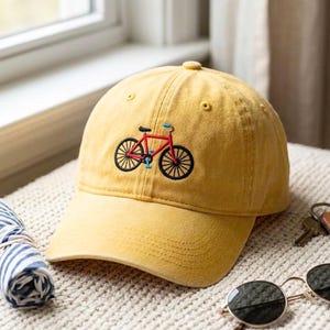 May include: A mustard yellow baseball cap featuring an embroidered graphic of a red bicycle with black wheels and blue accents. The cap has a structured crown, a curved brim, and stitched eyelets on the top panels for ventilation and durability.