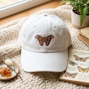 May include: A white baseball cap featuring a centered embroidered moth with brown and orange wings. The cap has a structured crown, a curved brim with visible stitching, and a button at the top. The design is clean and simple in its presentation.