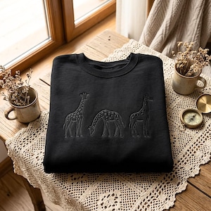 May include: A black crewneck sweatshirt featuring an embroidered design of three giraffes in the center. The embroidery is done in a dark thread, depicting one giraffe standing on the left, one grazing in the middle, and one standing on the right.