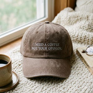 Pode incluir: Um boné de beisebol castanho com uma coroa estruturada e aba curva. A parte frontal apresenta um texto bordado a branco que diz: "I NEED A COFFEE NOT YOUR OPINION." O boné tem um design clássico de seis painéis com ilhós de ventilação.