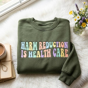 Op de afbeelding: Een olijfgroene sweater met ronde hals en het geborduurde tekst HARM REDUCTION IS HEALTH CARE in een veelkleurig, golvend lettertype. De letters zijn gekleurd in tinten lichtblauw, perzik, geel en lavendel op de donkere stof.