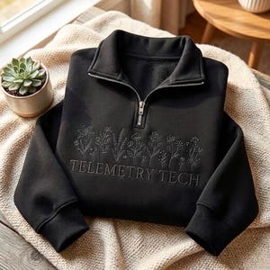 May include: A black quarter-zip sweatshirt featuring an embroidered line art design of various wildflowers across the chest. Below the floral pattern, the text TELEMETRY TECH is embroidered in a matching black thread. The garment has long sleeves.