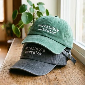 Puede incluir: Una gorra de béisbol verde apilada sobre una gorra de béisbol gris carbón. Ambas gorras presentan un acabado de tela desgastada y el texto bordado en blanco "unreliable narrator" en el panel frontal. Cada gorra tiene una visera curva.