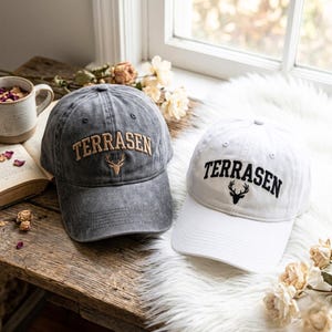May include: Two baseball caps, one in charcoal grey and one in white, both featuring the text TERRASEN in an arched font above a deer head icon. The grey cap has tan embroidery, while the white cap has black embroidery, both with a curved brim.