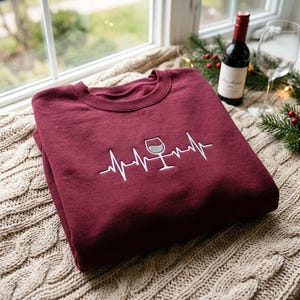 Puede incluir: Una sudadera de cuello redondo color granate con un diseño bordado en blanco de una línea de ritmo cardíaco que se transforma en la forma de una copa de vino. El bordado está centrado en el pecho de la prenda, hecha de tela lisa.